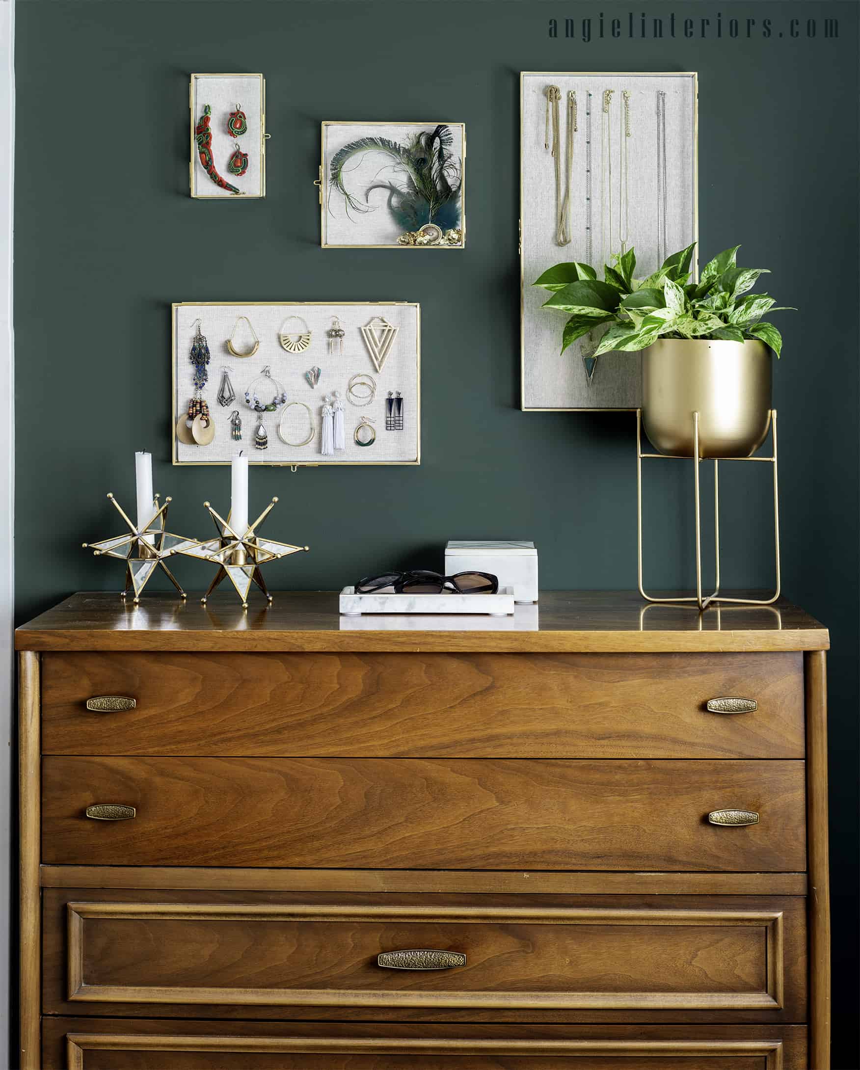 gold shadow boxes with jewelry hanging on a dark green wall over a wooden dresser