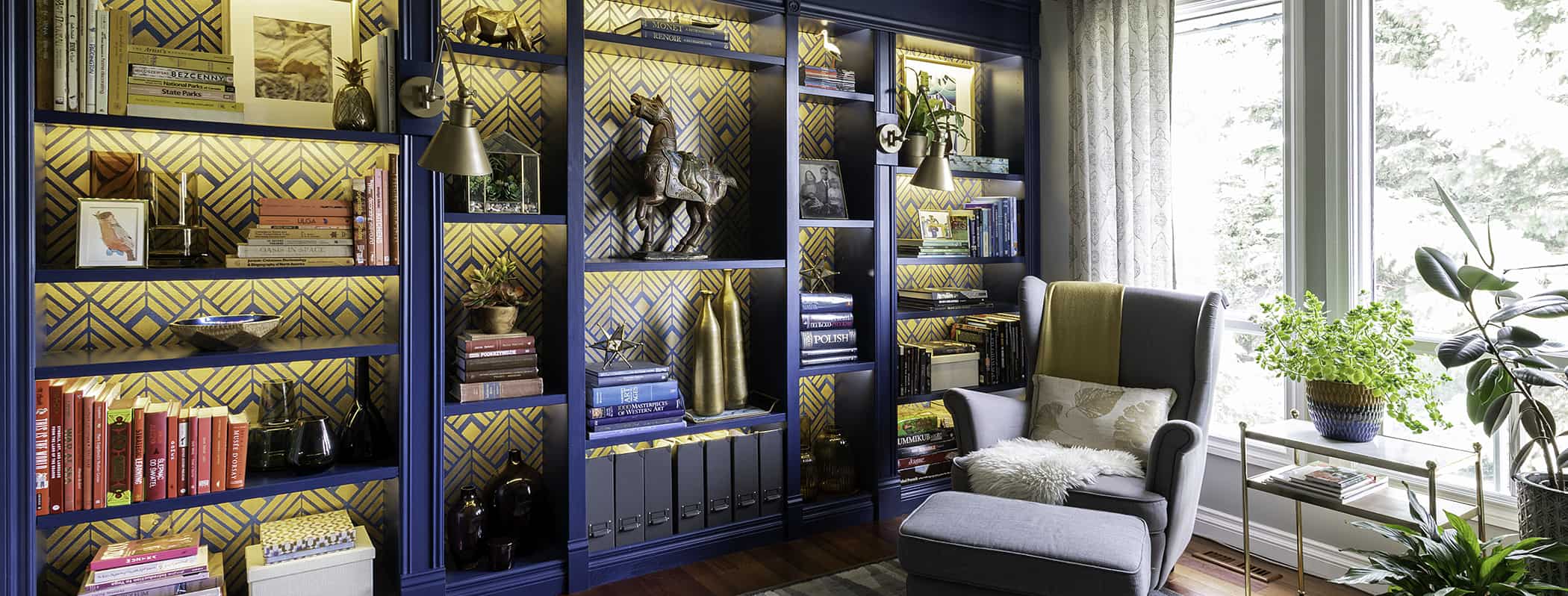 blue built in bookcase with gold stencil background and wingback armchair in front of a window