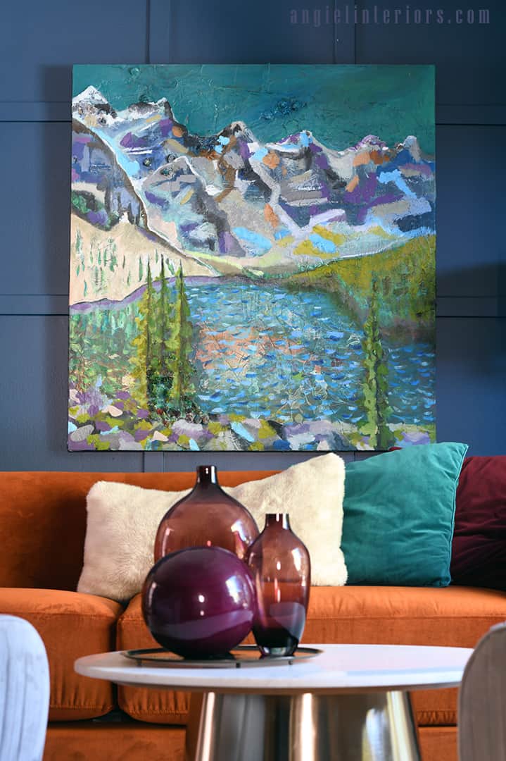 Painting of Moraine Lake on a blue grid wall above orange sofa with purple glass vases