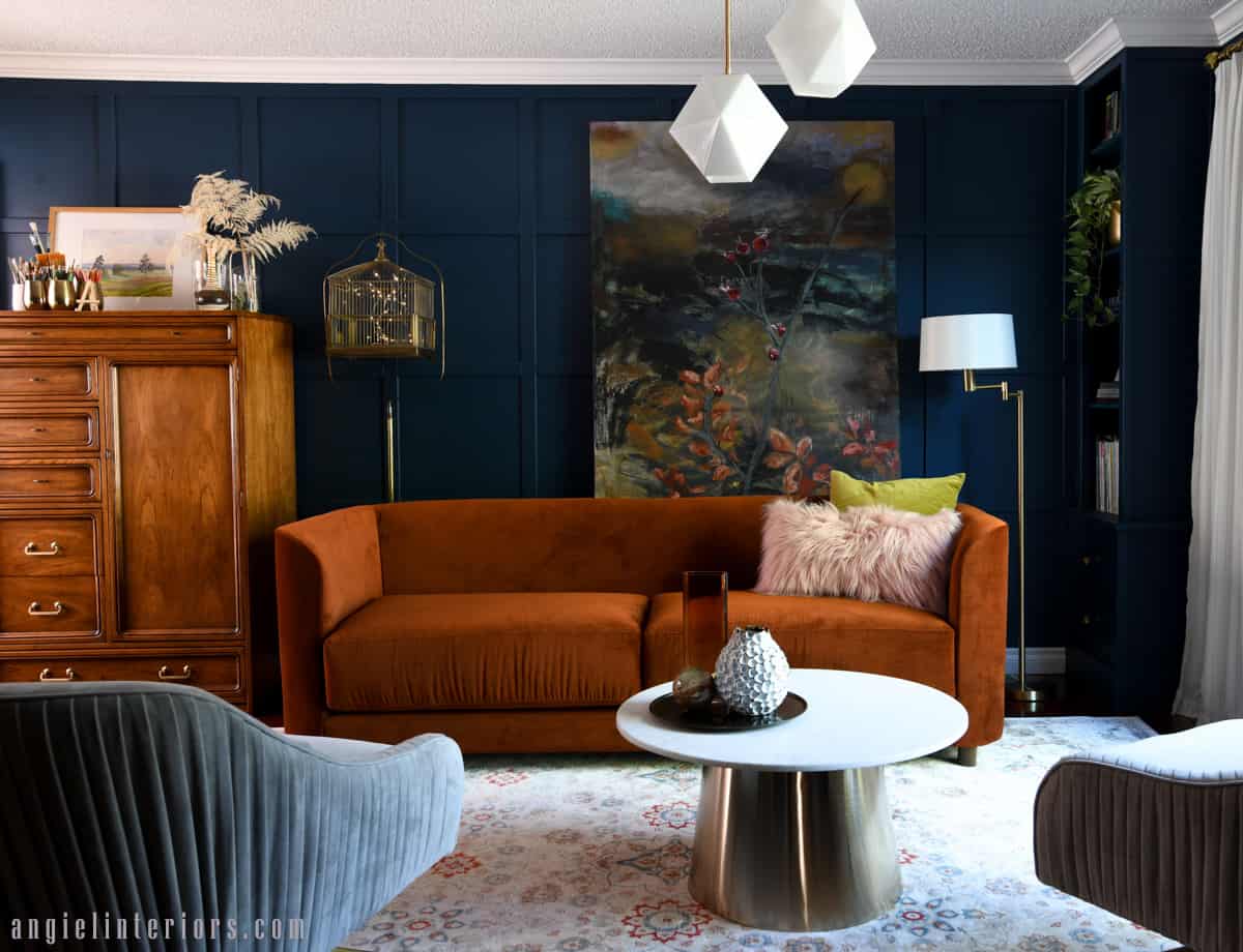 Living room with navy walls, orange sofa, abstract painting, marble and gold coffee table and velvet swivel chairs