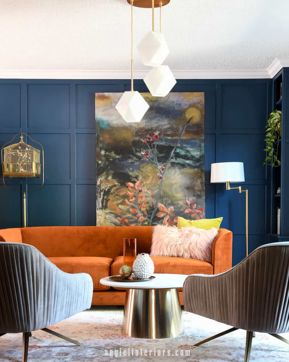 Living room with navy walls, orange sofa, abstract painting, marble and gold coffee table and velvet swivel chairs