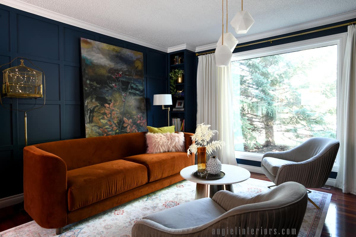 Living room with navy walls, built in bookcase, orange sofa, abstract painting, marble and gold coffee table and velvet swivel chairs