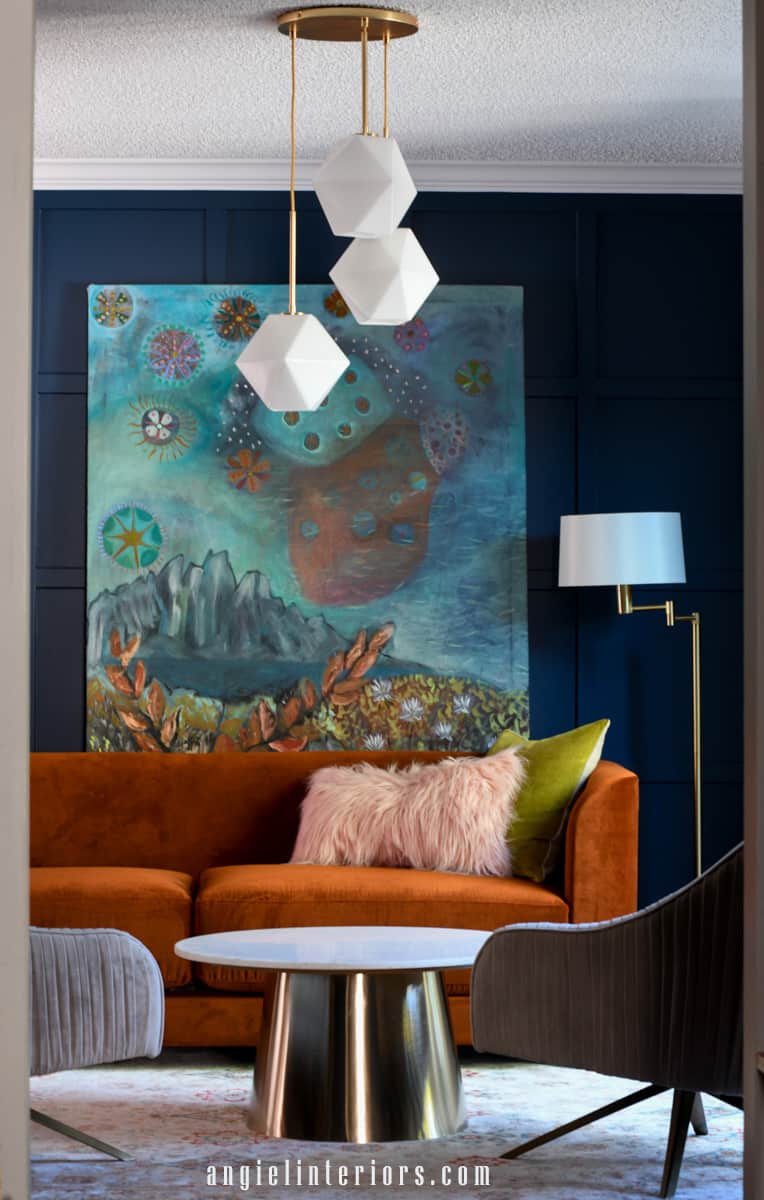 Living room with navy walls, orange sofa, abstract painting, marble and gold coffee table and velvet swivel chairs